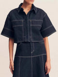 Midnight blue cropped short-sleeve contrast-stitch denim top, front view showing the pointed collar, button placket, chest patch pockets, and boxy cropped fit with bold white contrast stitching.