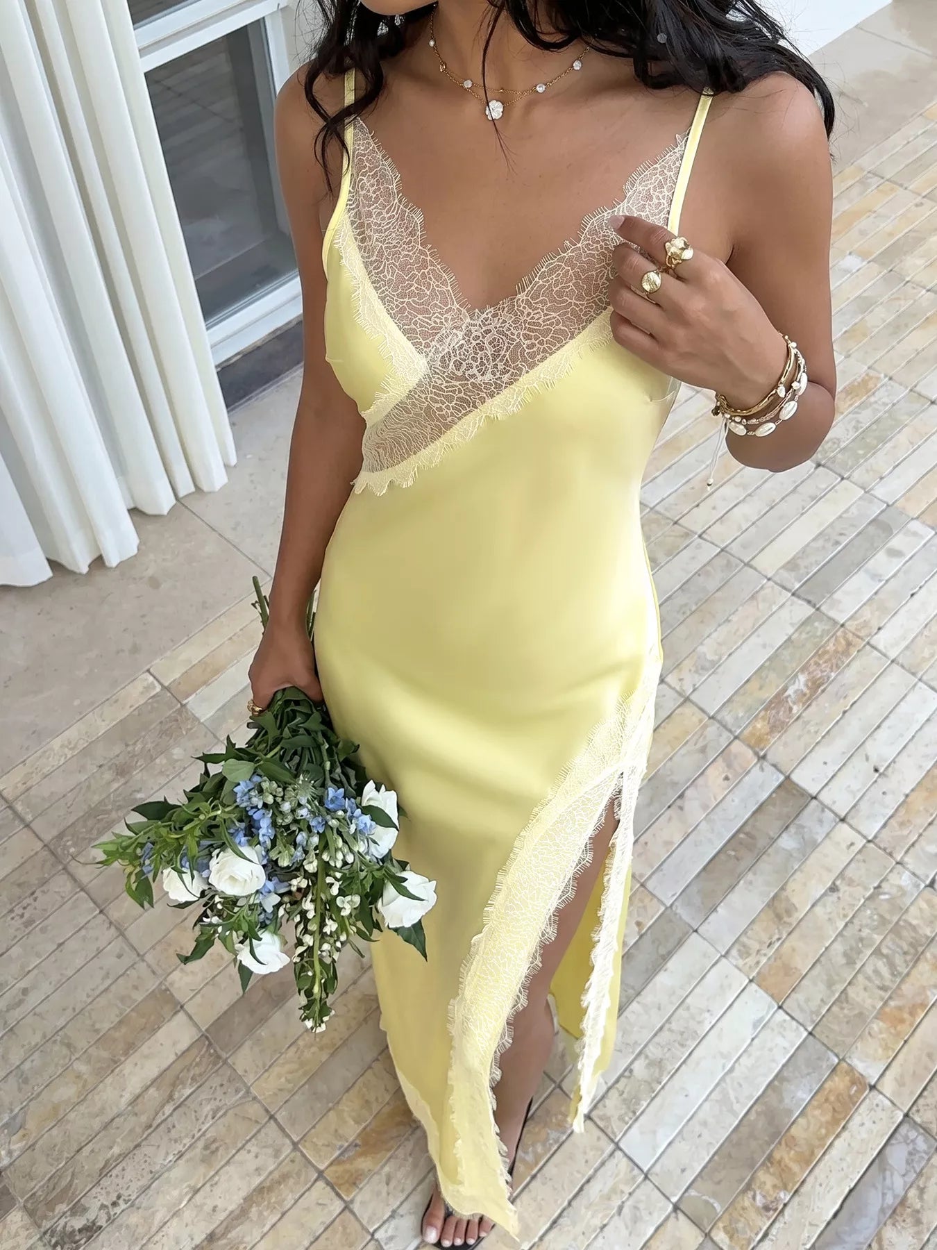 Model in the Pastel Yellow Lace-Trim Slip Midi Dress on an outdoor terrace, front view showing the white eyelash lace V-neckline, side slit, and lace hem with a white and blue bouquet.
