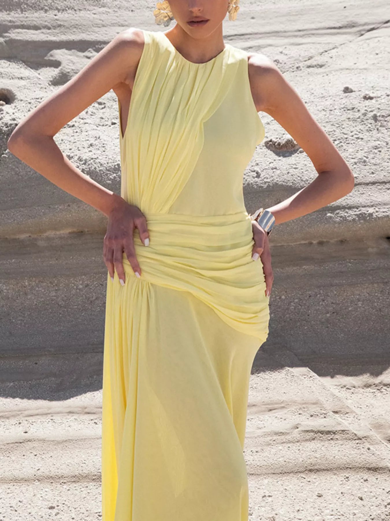 Mid-body view of the Pastel Yellow Sleeveless Ruched Maxi Dress, hands on hips on a beach, showing the asymmetric ruching gathered from bust through hip with a gold cuff bracelet.