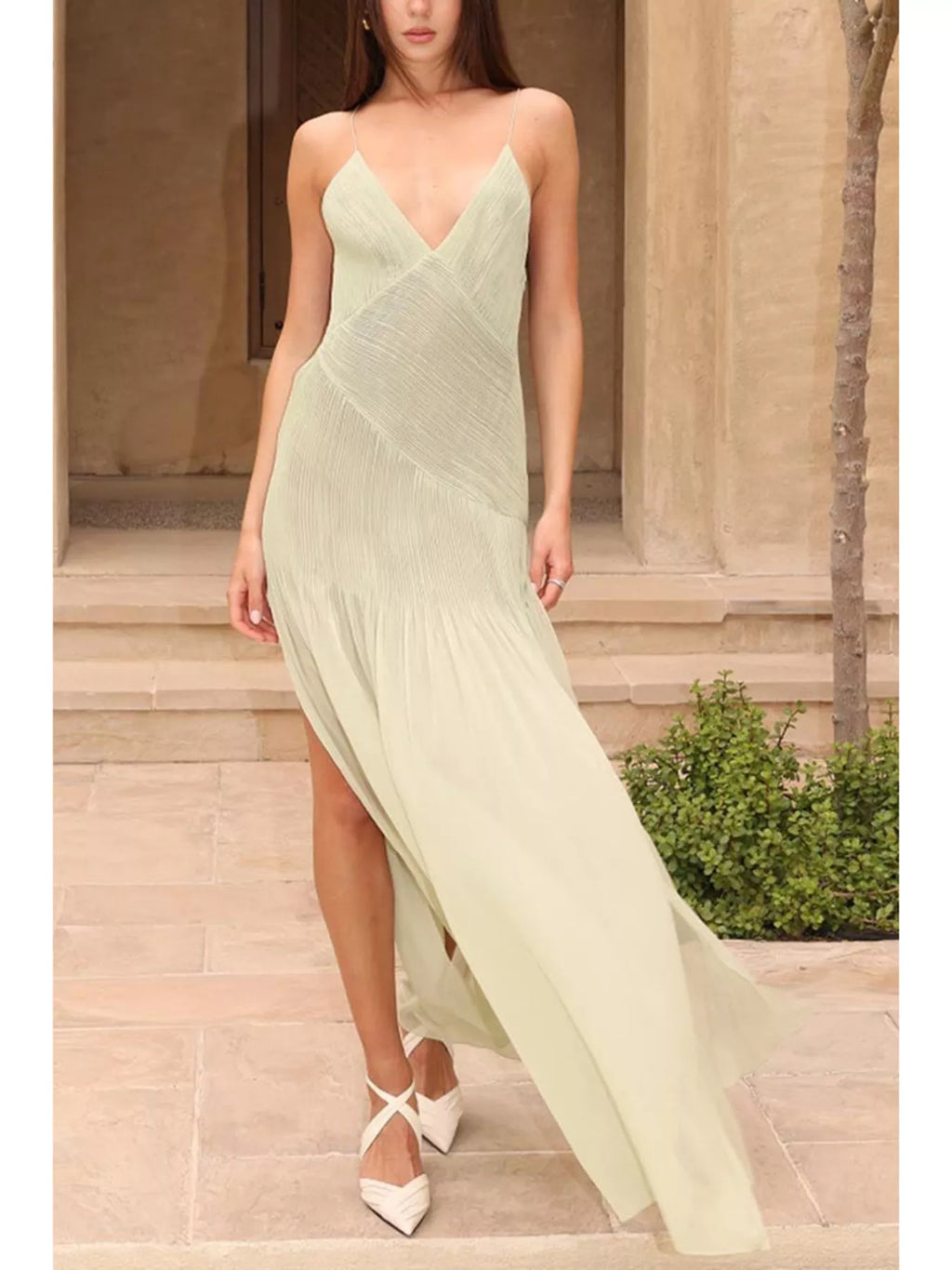 Sage green plissé maxi dress with deep V-neckline, wrap-style front, delicate spaghetti straps, and flowing pleated skirt, styled in a warm stone archway setting, full view walking.