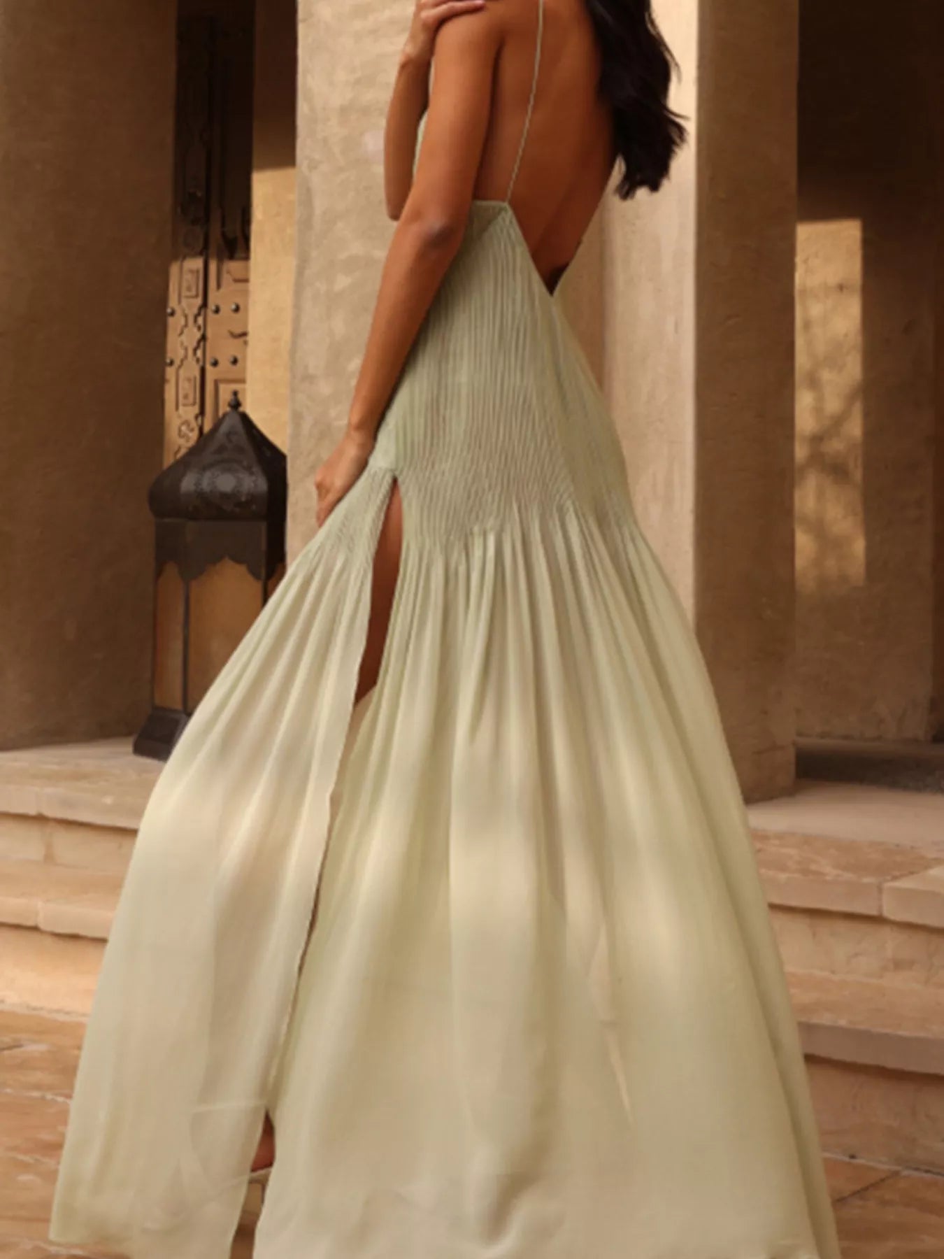 Three-quarter back view of a sage green plissé maxi dress, highlighting the open low back, delicate spaghetti straps, and fine vertical pleating, photographed poolside in warm natural light.
